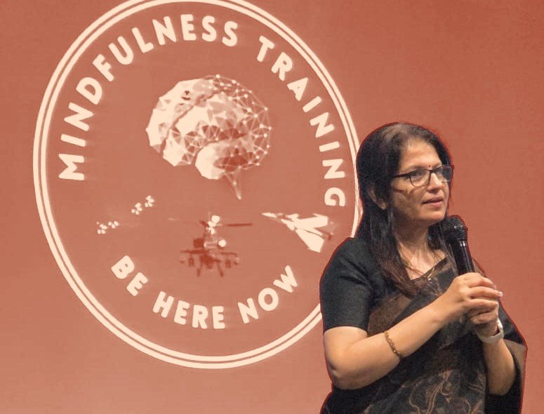 Shoba Shekar speaking in front of a Mindfulness Training, Be Here Now slide